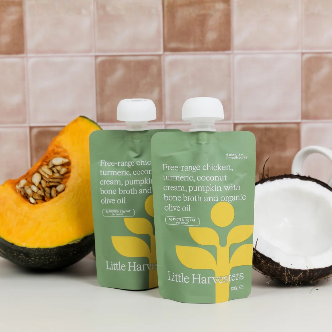 Two pouches of Little Harvester baby food stand on a white surface with a tiled background, flanked by a sliced pumpkin and a coconut half.